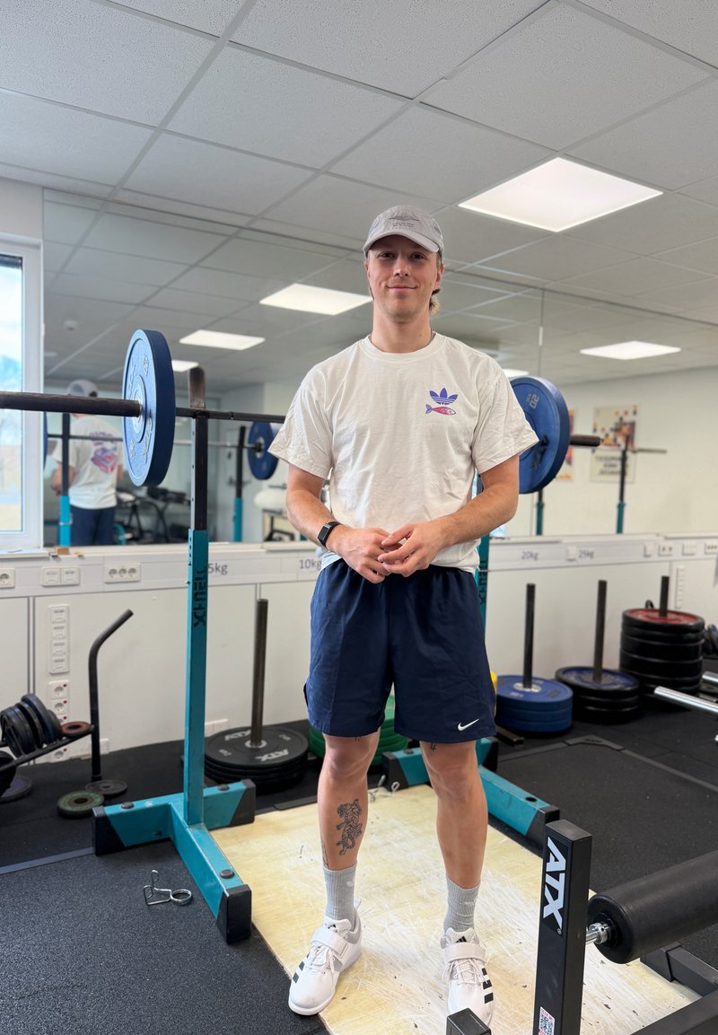 Young man in a white cap, white t-shirt, and blue shorts standing in a gym near a barbell rack with blue weight plates.