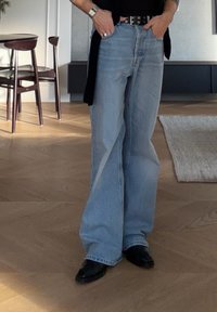 Άτομο που φοράει ανοιχτό μπλε τζιν παντελόνι με φαρδιά μπατζάκια, μαύρα παπούτσια και μαύρη ζώνη, σε φωτεινό δωμάτιο με ξύλινο πάτωμα και κοντινές καρέκλες τραπεζαρίας.