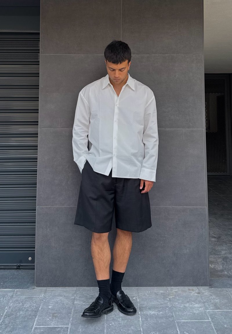 Chemise blanche à manches longues et boutons, shorts noirs ajustés, chaussettes noires et mocassins noirs. Se tenant devant un mur en carrelage gris.