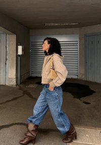 Beige cropped hette, løse blå jeans og brune hæle støvler. Outfittet har en teksturert skulderveske og krøllete hår. Moderne urban bakgrunn.