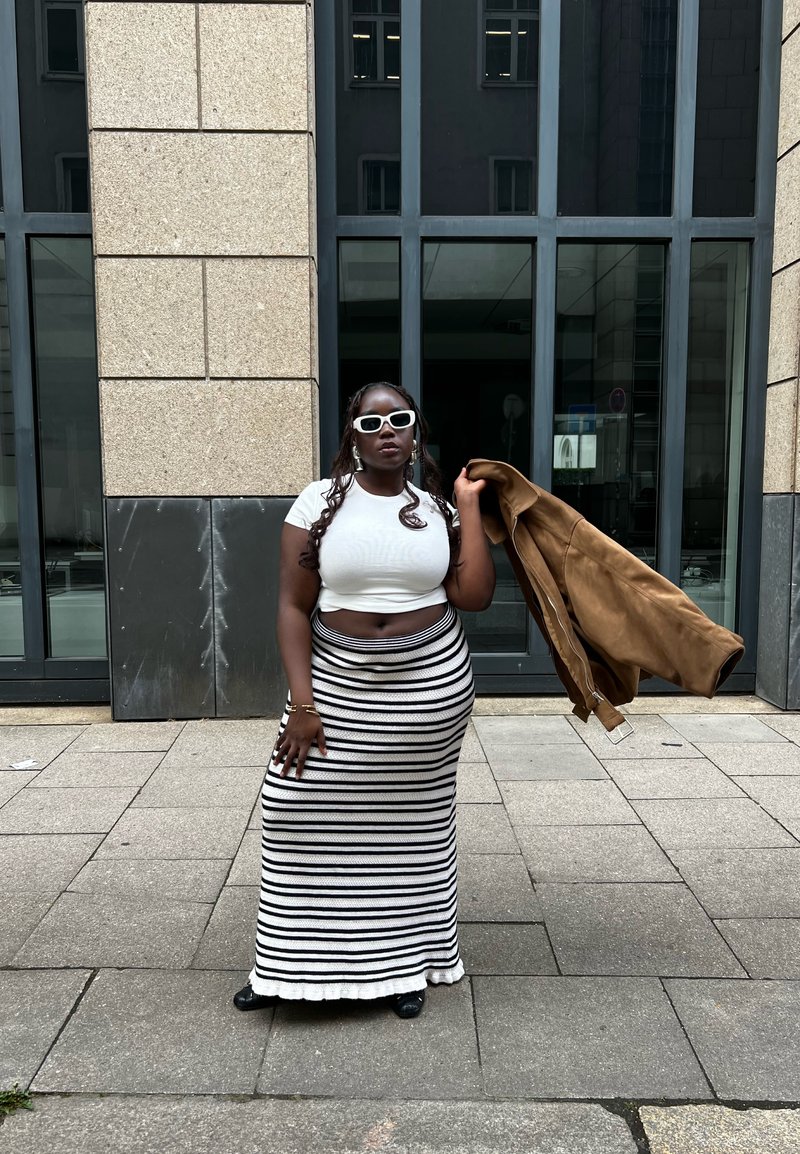 Femme portant un crop top blanc, une jupe longue rayée noire et blanche, des lunettes de soleil blanches, tenant une veste marron, debout sur un trottoir en ville.