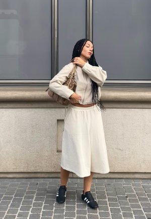 Chaqueta corta beige ligera con mangas largas, falda midi crema y zapatillas deportivas negras con rayas blancas; sosteniendo un bolso texturizado.