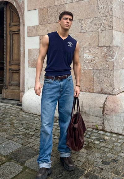 Top sin mangas navy, jeans de color azul claro, cinturón de cuero marrón, bolsa marrón oscuro y botas. El fondo presenta una pared de piedra y una puerta de madera.