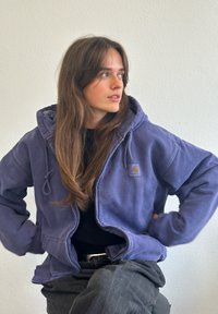 Fialová mikina s kapucňou v uvoľnenom strihu, so zipsom, šnúrkami a logom, skombinovaná s čiernym topom a sivými nohavicami.
