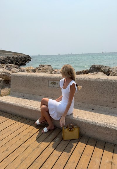 Vestido de ganchillo blanco con dobladillo ondulado, sandalias planas blancas y un bolso de paja amarillo con un asa circular, sentado en un banco de piedra.