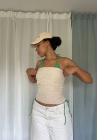 Femeie ajustând un top bej fără bretele peste un bikini verde cu guler, purtând pantaloni albi și o șapcă crem, stând în fața unor draperii transparente.