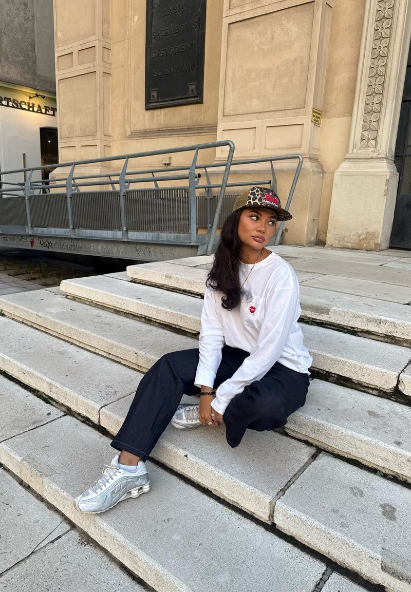 Giovane adulto seduto sui gradini di pietra, indossando una maglietta bianca a maniche lunghe con un cuore rosso, pantaloni neri a gamba larga e sneaker argentate.