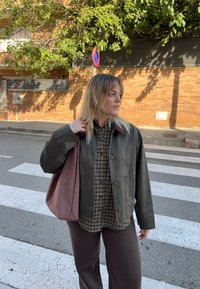 Giacca verde scuro con due tasche frontali sopra una camicia a quadri marrone e beige, abbinata a pantaloni marroni e una borsa a tracolla color malva.