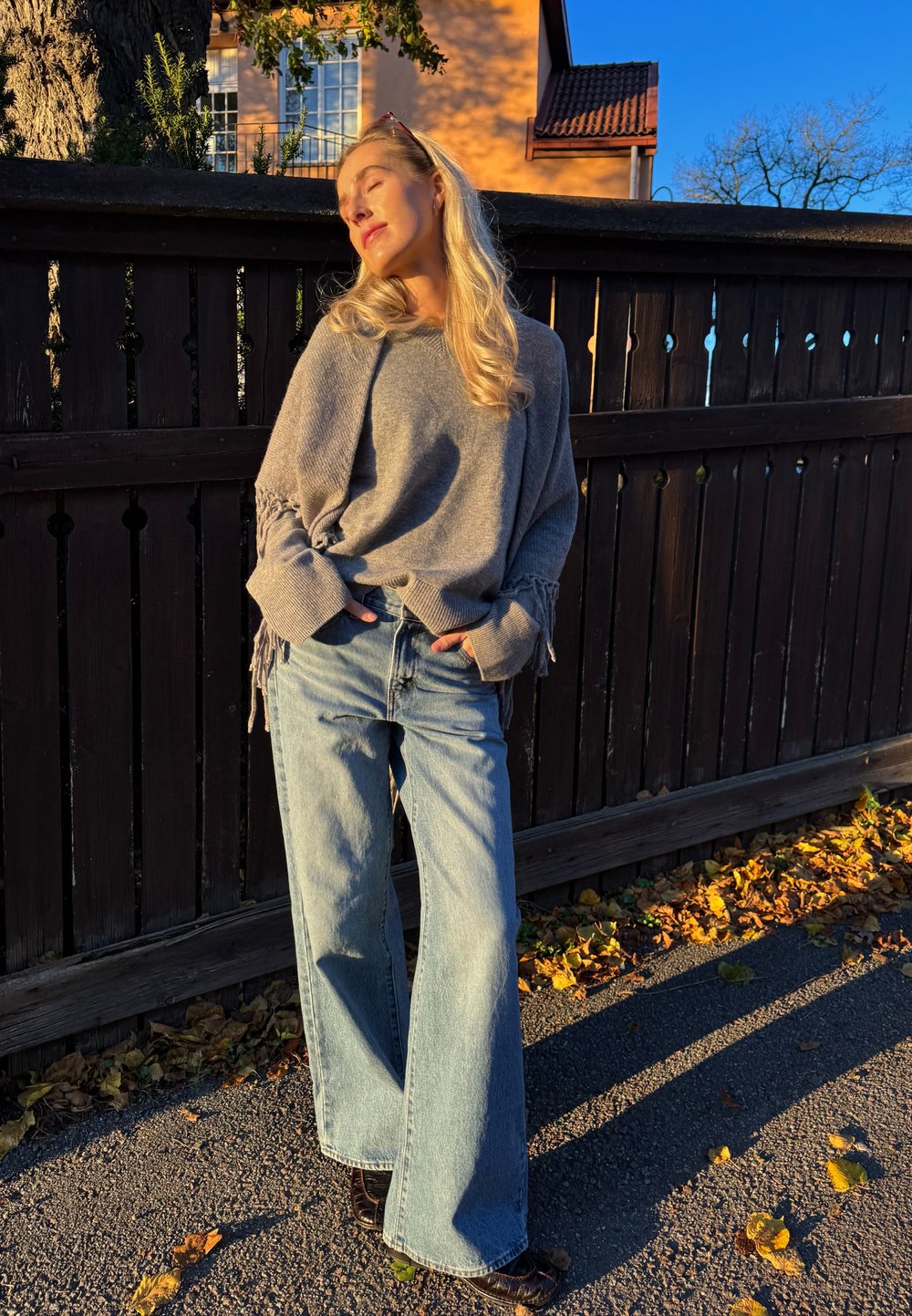 Pull gris surdimensionné avec détails en franges, associé à un jean ample bleu clair. Le décor comprend une clôture en bois et des feuilles d'automne.