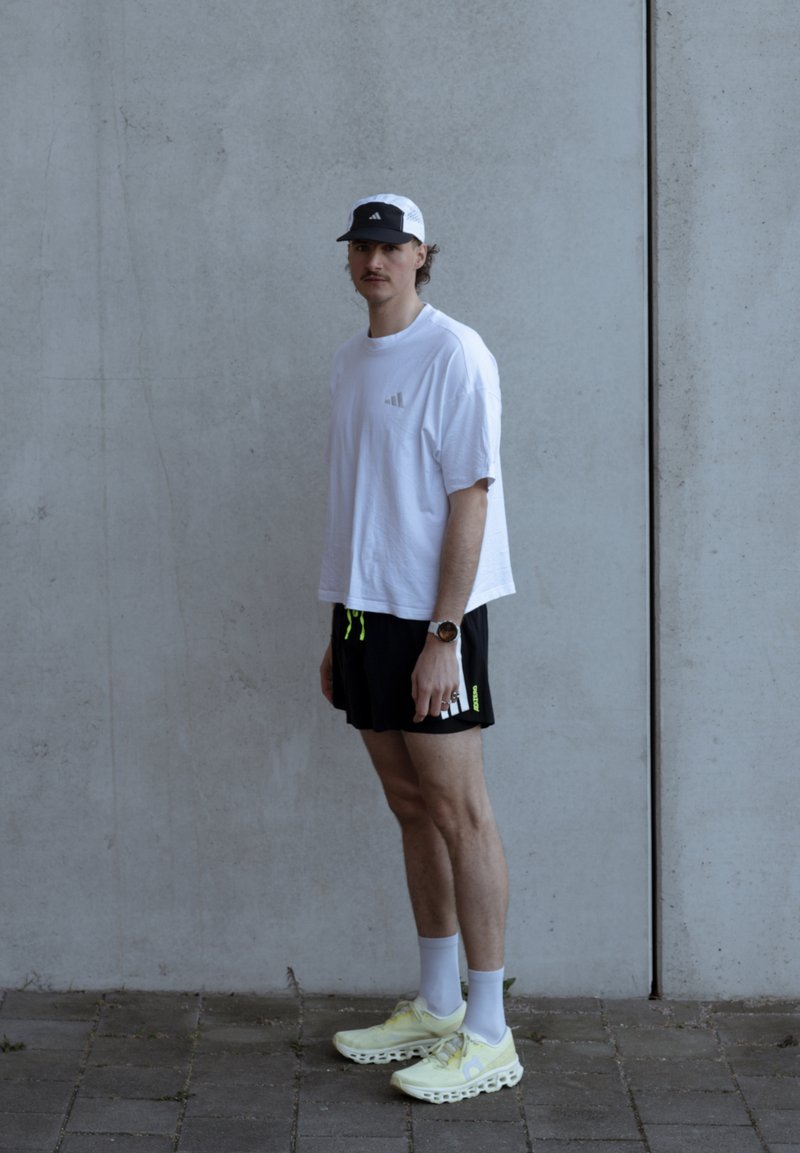 Ung mand iført hvid kasket, oversized hvid Adidas T-shirt, sorte shorts, hvide sokker og lysegule sneakers, der står op ad en grå betonvæg.