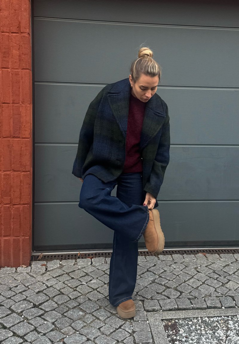 Temen tartan volnen plašč, bordo pulover, široke modre denim hlače in bež semiš gležnjarji; stoji na tlakovanem tlakovcu blizu sivega ozadja.