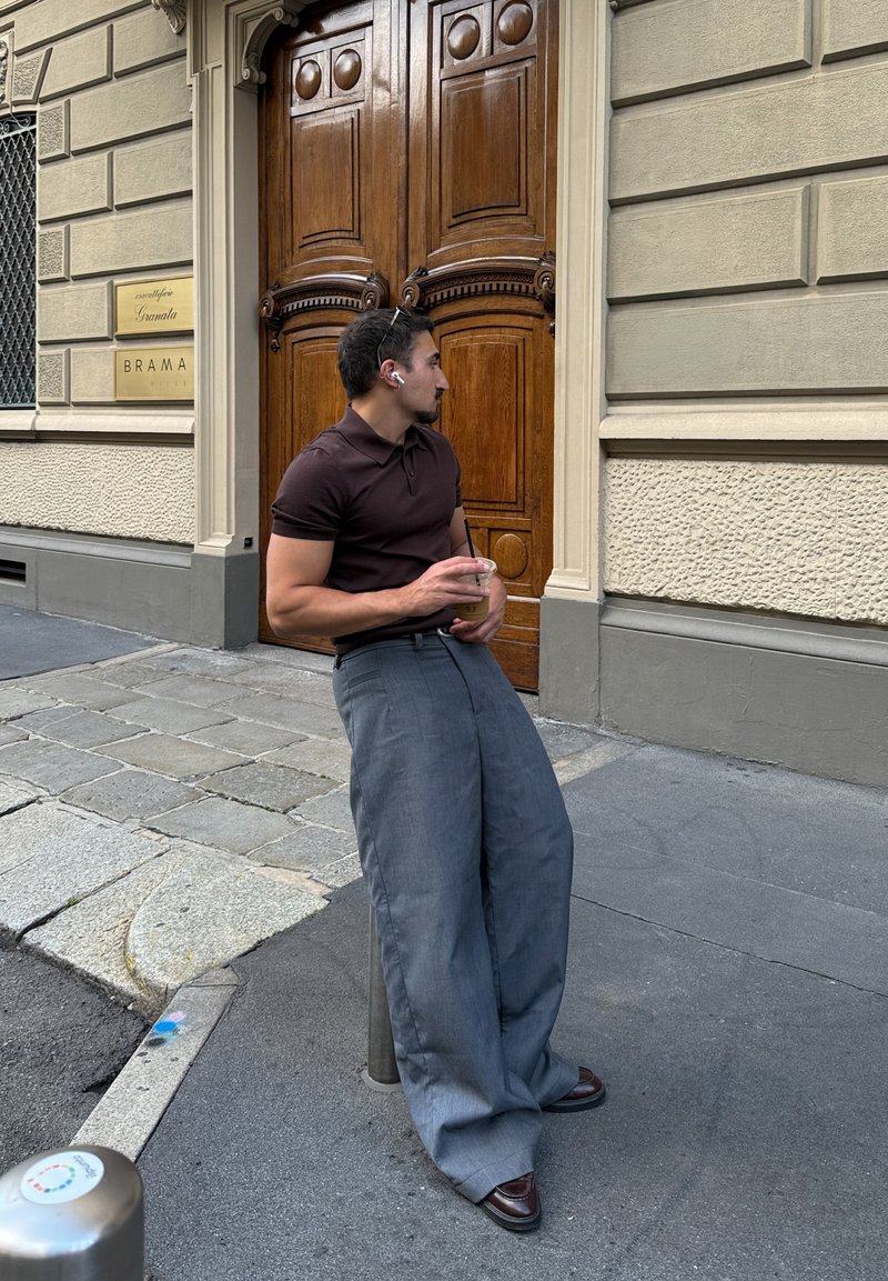 Homme en chemise sombre et pantalon gris large appuyé sur un poteau tenant un café glacé près de grandes portes doubles en bois sur le trottoir de la ville.