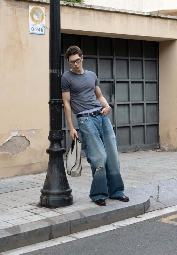 Joven con gafas, camiseta gris y vaqueros anchos se apoya en un farol negro sosteniendo una bolsa beige en una acera de la ciudad.