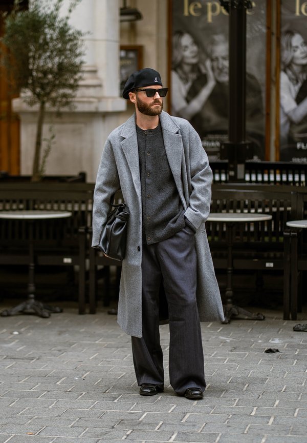 Un hombre barbudo con gafas de sol y una boina negra está de pie frente a una cafetería, vestido con un abrigo gris y con un bolso de cuero negro.