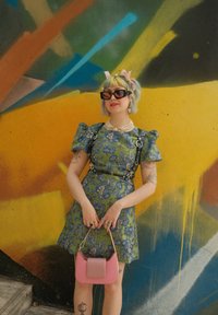 Πράσινο λουλουδάτο φόρεμα με φουσκωτά μανίκια, μαύρος ιμάντας και ροζ τσάντα. Σε φόντο πολύχρωμου αφηρημένου τοιχογραφίας.