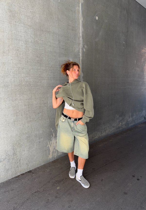 Jeune personne portant un short oversize, un sweat à capuche court et des chaussures à enfiler à carreaux, debout contre un mur en béton gris, écoutant des écouteurs.