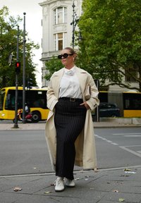 Palton bej, bluză albă, fustă neagră cu dungi, adidași albi. Ochelari de soare și o geantă tip plic completează ținuta. Fundal urban.