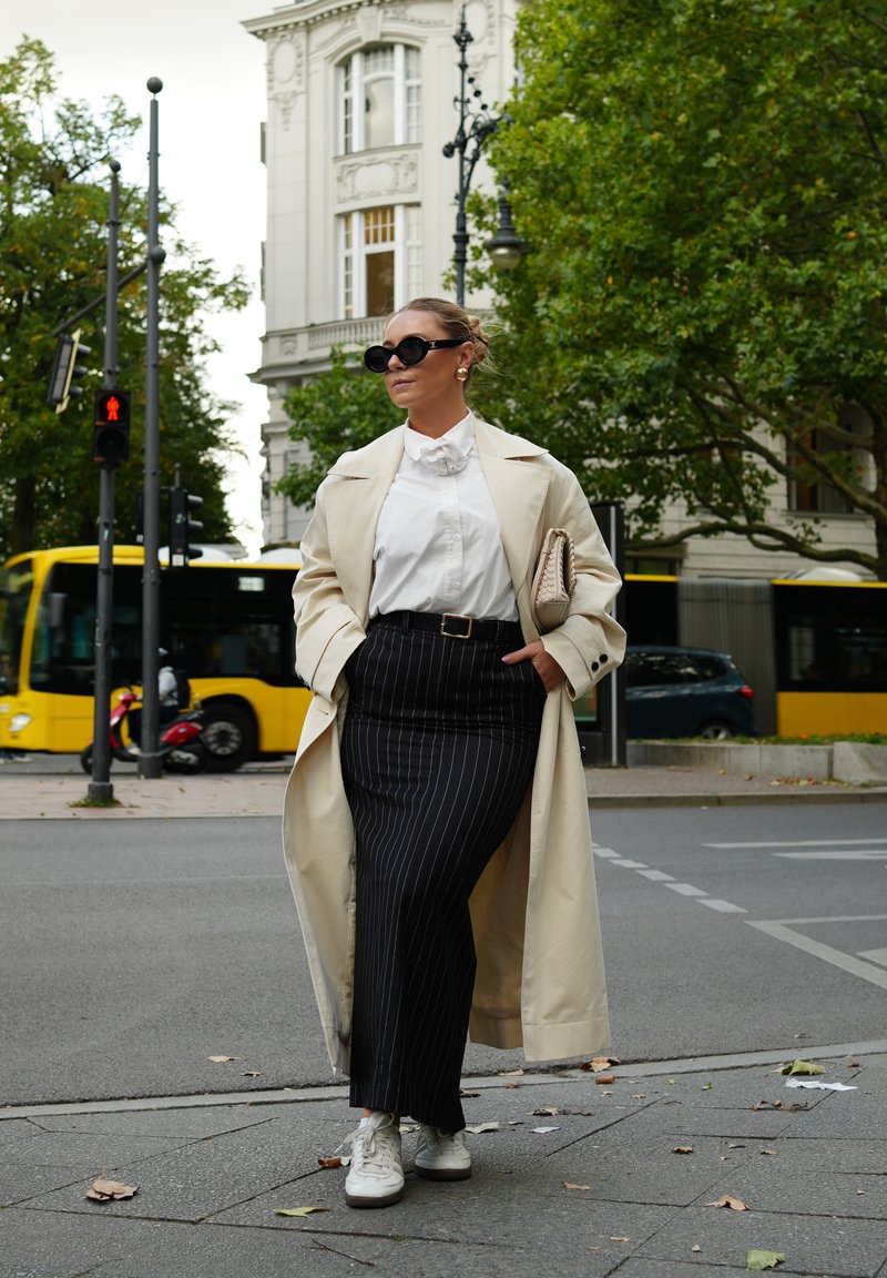 Manteau beige, blouse blanche, jupe noire à rayures, baskets blanches. Des lunettes de soleil et une pochette complètent la tenue. Fond urbain.