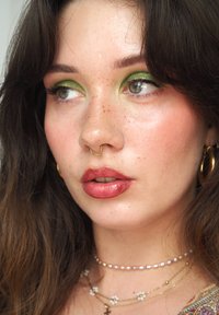 Nahaufnahme eines Gesichts mit grünem Lidschatten, glänzenden Lippen, einem goldenen Septumring und geschichteten Halsketten mit Perlen und Blumen. Mit Sommersprossen.