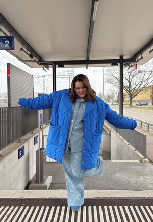 Abrigo acolchado azul, de gran tamaño, con grandes bolsillos, que se lleva sobre un atuendo azul claro con detalles brillantes. El escenario es una plataforma de estación de tren.