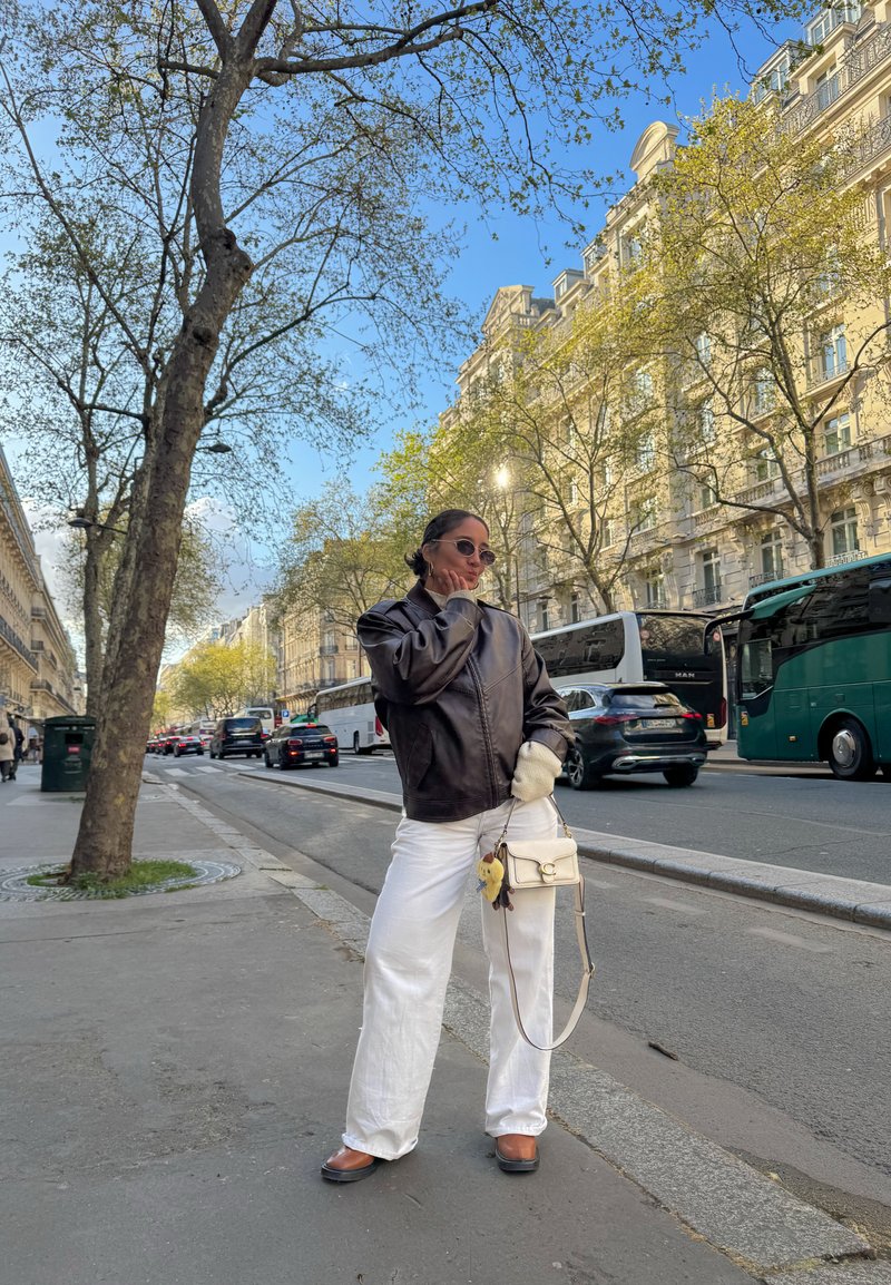 Femme en veste noire, pantalon blanc et lunettes de soleil posant sur le trottoir de la ville avec des arbres, des voitures et des bâtiments historiques en plein jour lumineux.
