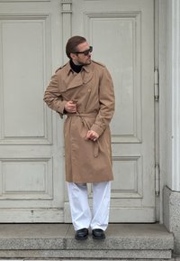 Trench-coat beige à taille ceinturée, boutonnage croisé et large col, associé à un pantalon blanc et des chaussures noires. Debout sur des marches.