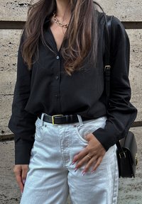 Sort knap-til-krave skjorte med langeærmer, parret med højtaljede hvide denim jeans og et sort bælte. Guld kæde halskæde synligt.