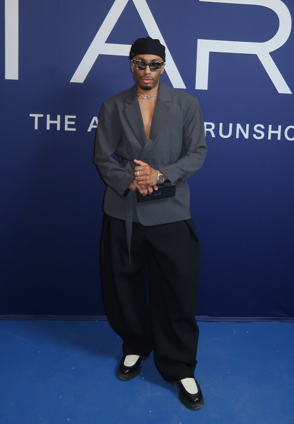 Hombre con gorro negro, blazer gris abierto, pantalones negros anchos, zapatos blancos y negros, gafas de sol y joyas, de pie frente a una pared azul con texto.