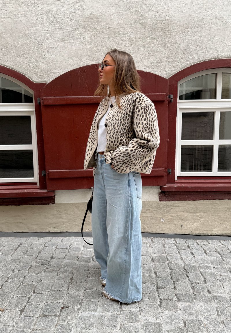 Jeune femme aux longs cheveux portant des lunettes de soleil, une veste à motif léopard, un haut blanc, un jean à jambes larges, tenant un petit sac à main noir dans une rue pavée.