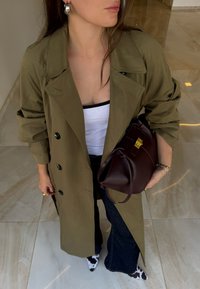 Olíva zöld trench coat fekete gombokkal, fehér felső fölött. Sötét farmer, bővülő nadrág és bordó kézitáska arany kiegészítőkkel.