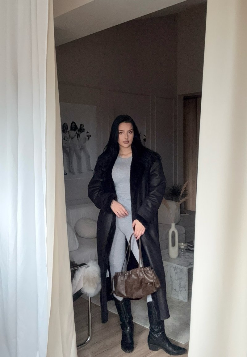Schwarzer langer Mantel mit Fellkragen, graues enges Outfit, braune Handtasche und schwarze kniehohe Stiefel. Innenraum mit neutralen Farben.
