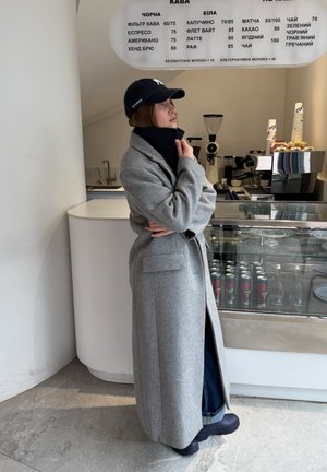 Persona con un abrigo largo gris y una gorra negra está dentro de una cafetería moderna, mirando un menú sobre el mostrador con opciones de café y bebidas.