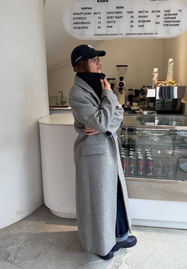 Persona con un lungo cappotto grigio e un berretto nero si trova all'interno di un bar moderno, guardando un menù sopra il bancone con opzioni di caffè e bevande.