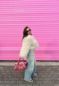 Bílá chlupatá bunda, světle modré široké džíny a růžová kabelka. Černé sluneční brýle doplňují vzhled na pozadí s růžovými pruhy.