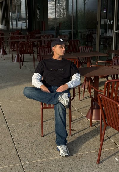 Joven con gafas, gorra negra y camisa de capas sentado con las piernas cruzadas en una silla de una cafetería al aire libre vacía con mesas y sillas rojas.