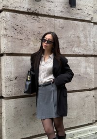 Veste noire ample, pull en maille torsadée beige, jupe plissée grise, sac à main noir avec des accents dorés, lunettes de soleil noires, et collants noirs transparents.