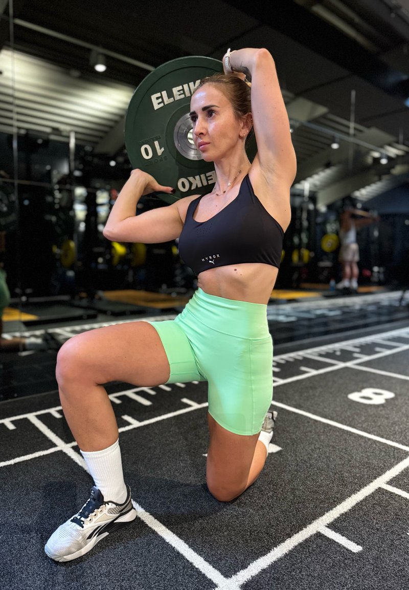 Femme en brassière noire et short vert agenouillée tenant une plaque d'haltère derrière la tête dans une salle de sport.