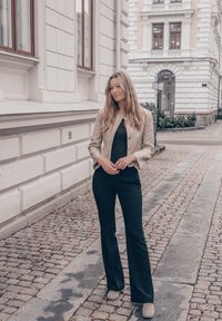Jovem mulher com cabelo longo vestindo uma jaqueta bege e calças pretas está em uma rua de paralelepípedo entre edifícios clássicos.