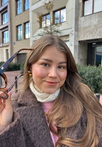 Des lunettes de soleil en écaille de tortue tenues par une personne, avec un design à bras courbé. La personne porte une écharpe tricotée blanche et un manteau gris.