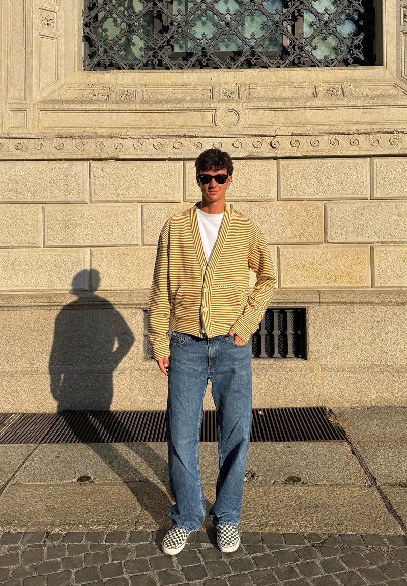 Gul- och vitrandig kofta med v-ringning, kombinerad med ljusblå jeans och rutiga slip-on skor. Kraftiga skuggor på trottoaren.