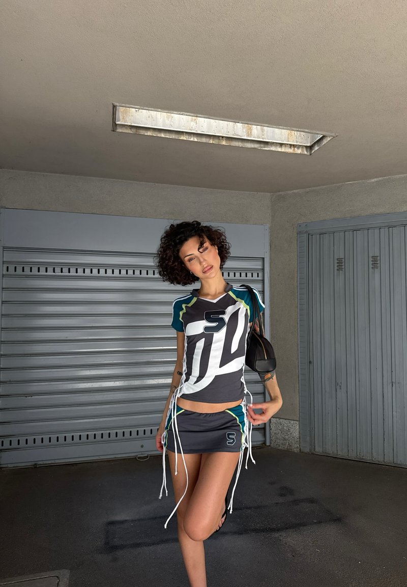 Ung person med krøllet hår iført et sporty todelt sæt, der poserer i en garage med lukkede metal døre og en loftslygte over.