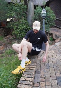 Jeune homme portant une casquette et une tenue de sport assis sur un mur en briques bas, ajustant ses baskets jaunes près d'un lampadaire de jardin et de chemin.