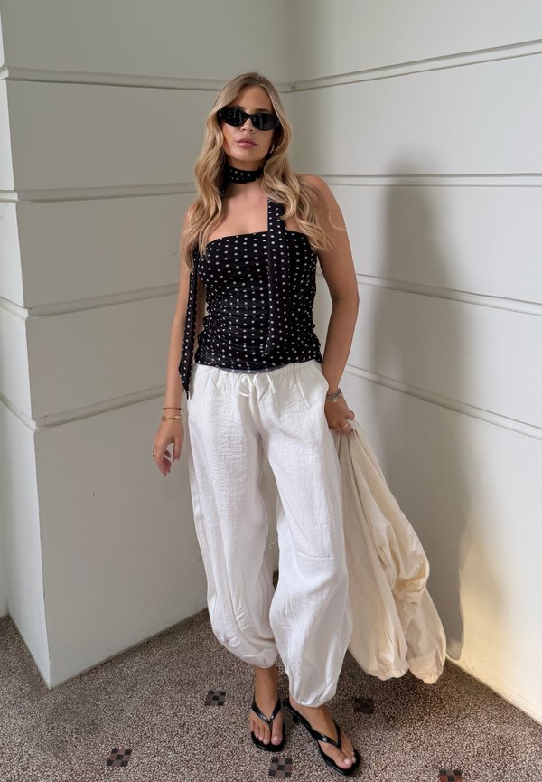 Woman wearing black polka dot top, white loose pants, black sandals, and sunglasses, standing against white wall holding beige jacket.