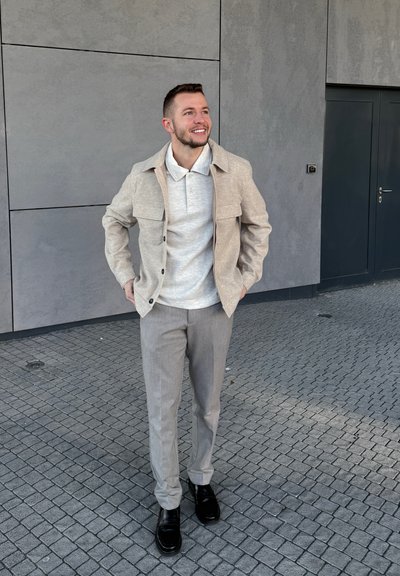 Hombre sonriente con chaqueta beige, polo gris claro, pantalones grises y zapatos negros de pie sobre un pavimento de adoquines delante de una pared gris y una puerta.