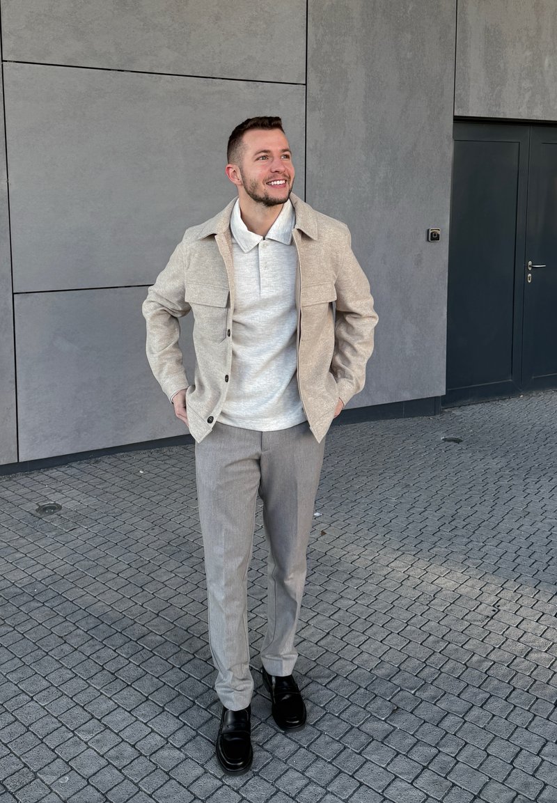 Smiling man wearing a beige jacket, light grey polo shirt, grey trousers, and black shoes standing on cobblestone pavement in front of a grey wall and door.