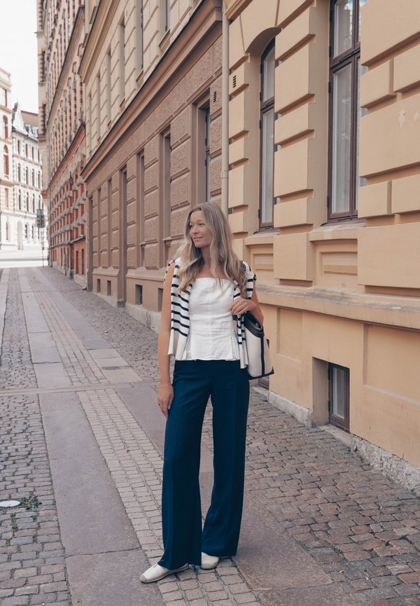 Kobieta z długimi blond włosami stoi na brukowanej ulicy obok beżowego budynku, ubrana w biały top, granatowe spodnie i paskowany sweter narzucony na ramiona.