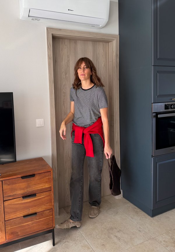 Une femme se tient dans l’encadrement de la porte d’une cuisine moderne, un sac à main foncé à la main. Elle porte une chemise grise, un jean et un pull rouge noué autour de la taille.