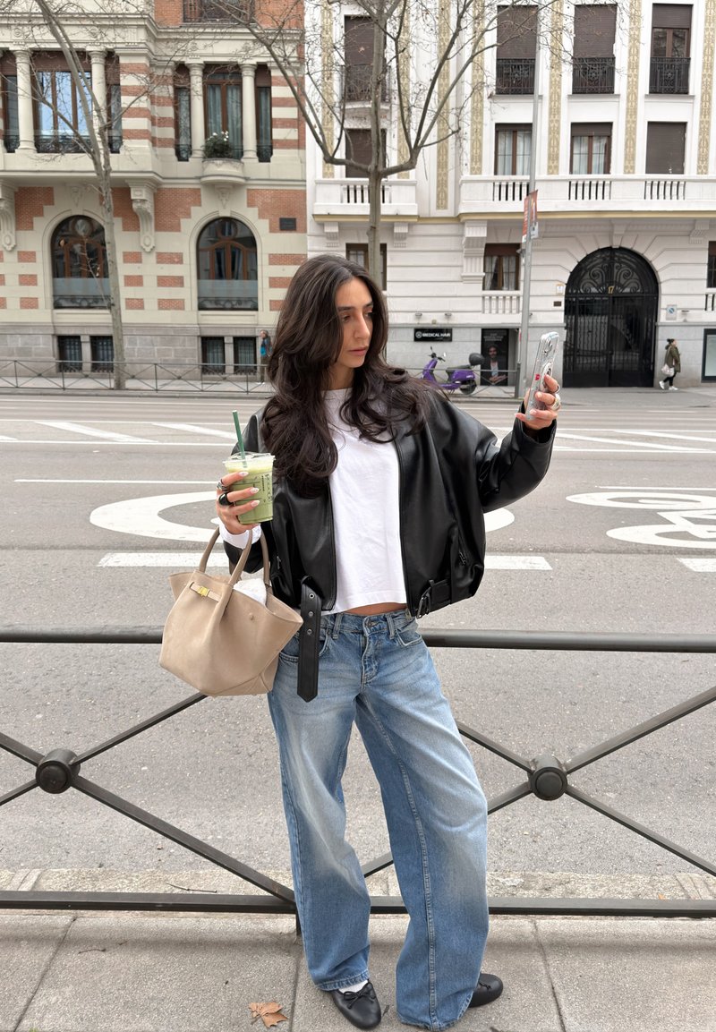 Femme aux longs cheveux foncés portant une veste en cuir noire et un jean bleu, tenant un verre vert et prenant un selfie dans une rue de la ville.