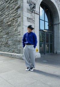Blå sweater, grå plisserede brede bukser, sorte sneakers med turkise detaljer, sort kasket og solbriller. Baggrund med stenmur.