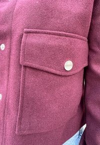 Burgundi vörös gyapjú dzseki, filces textúrával, mellzsebből és ezüst színű patentgombokból áll a záródáshoz. Közeli részlet a szövet mintázatáról.